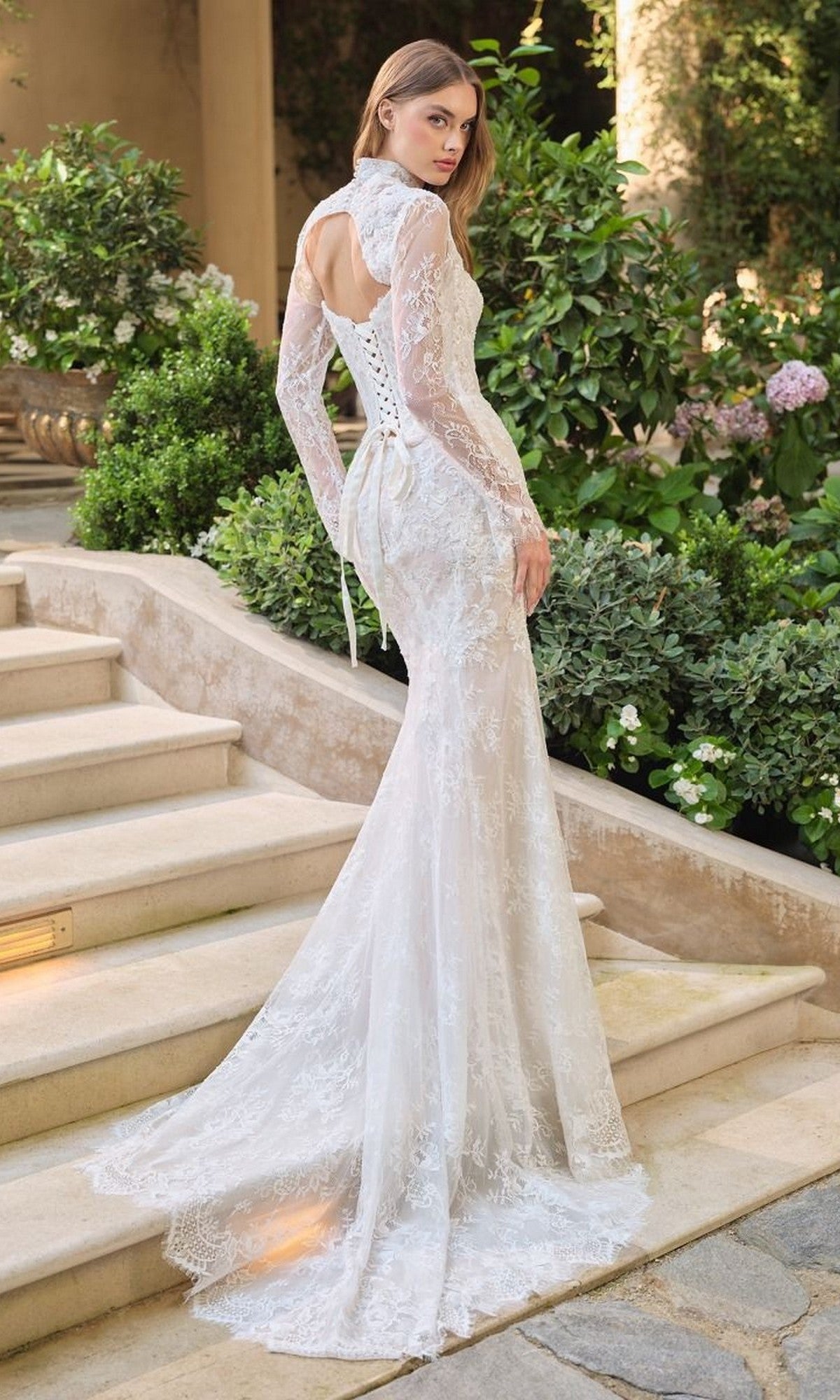 A woman stands on stone steps outdoors in the Andrea and Leo WL071 long wedding dress, featuring beaded lace, a detachable lace bolero, high neckline, open back, and long train. She is surrounded by lush greenery and flowers.